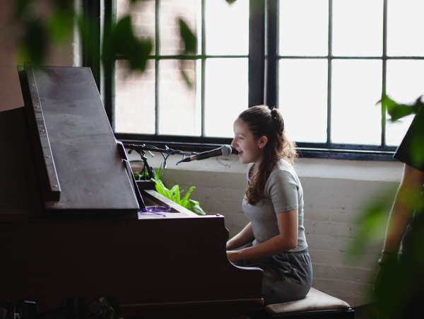 Desautels performing behind the baby grand piano at Dough Company, located in the Kilburn Mill. Desautels is the vocalist, pianist, and occasional guitarist and banjoist in her band, while her stepfather plays acoustic guitar.